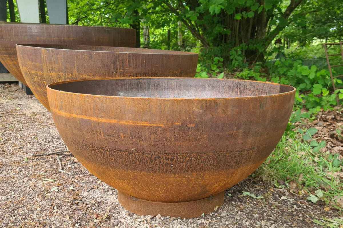 Large rusted Corten steel water bowls arranged outdoors on gravel with green trees and shrubs in the background.