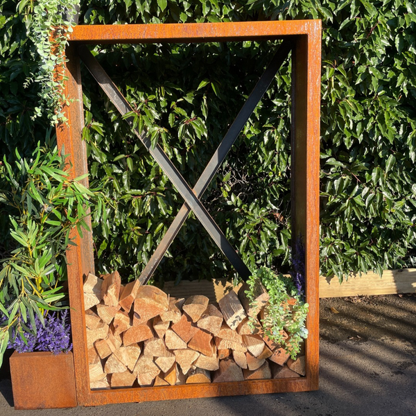 Corten Steel firewood holder with stacked firewood against a green hedge.
