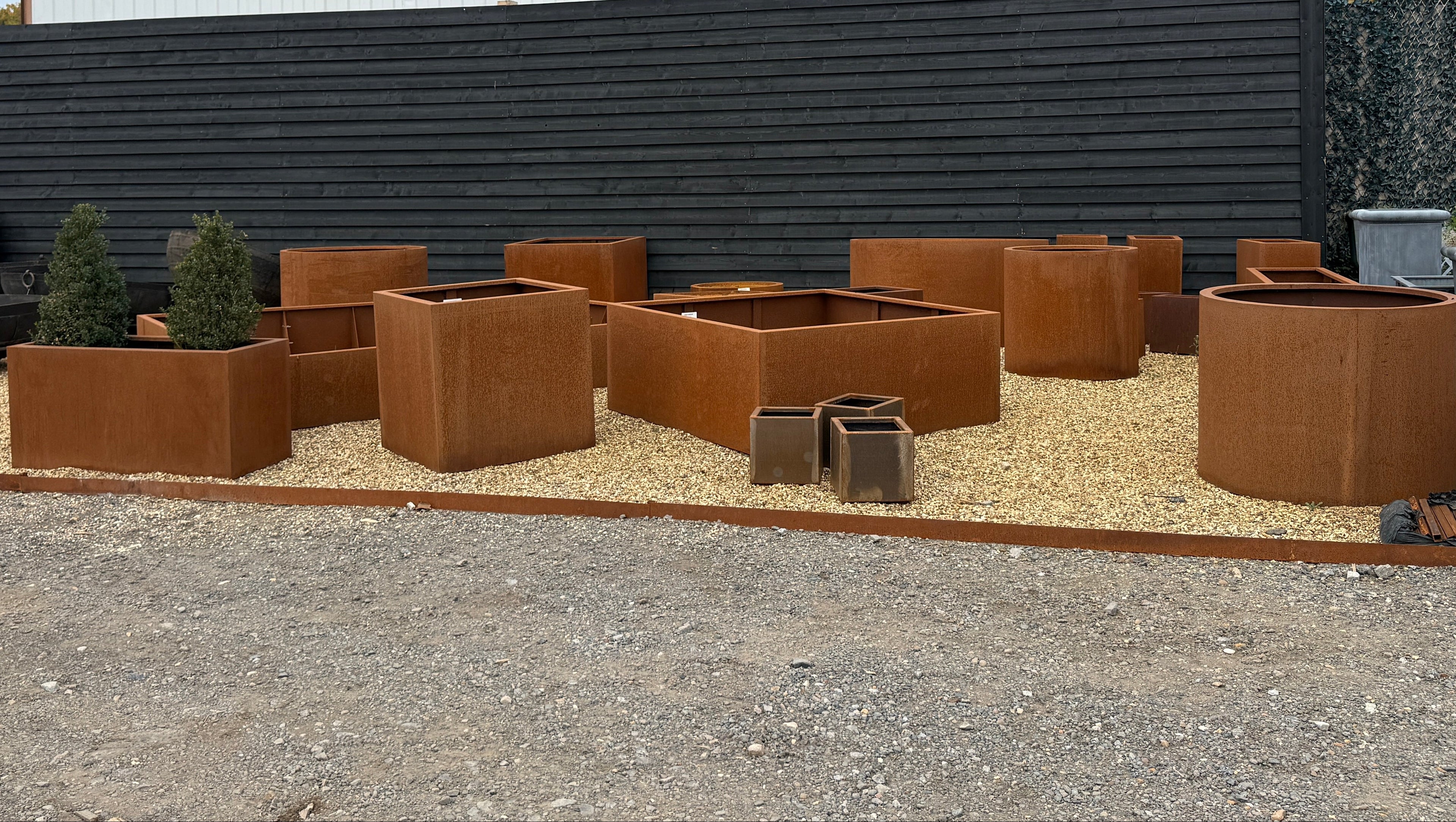 Collection of rust-colored metal planters and fire pits on a gravel surface with a building in the background.