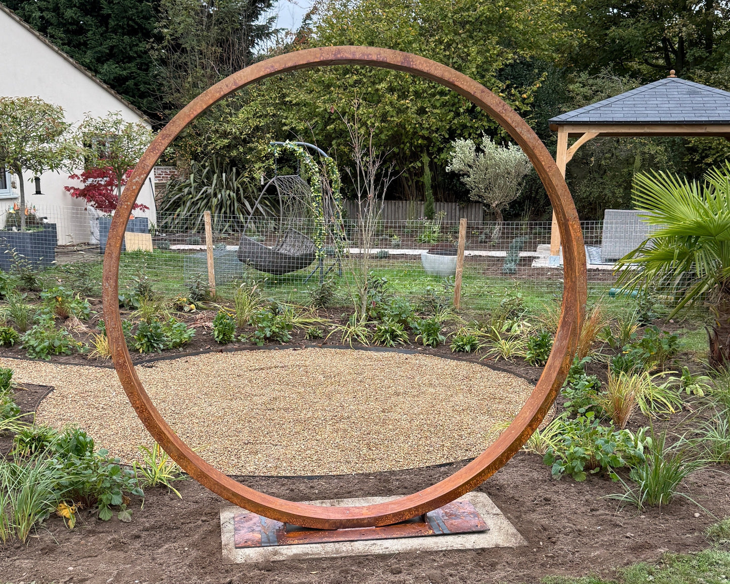 Large corten metal circular moongate sculpture in a garden setting with trees and a gazebo in the background.