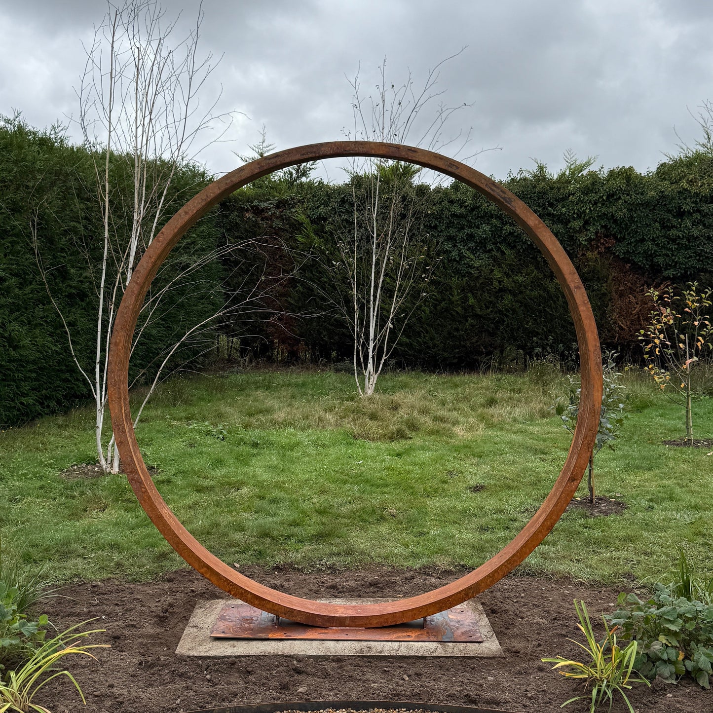 Rust-colored circular corten metal moongate sculpture in a garden setting with a cloudy sky.