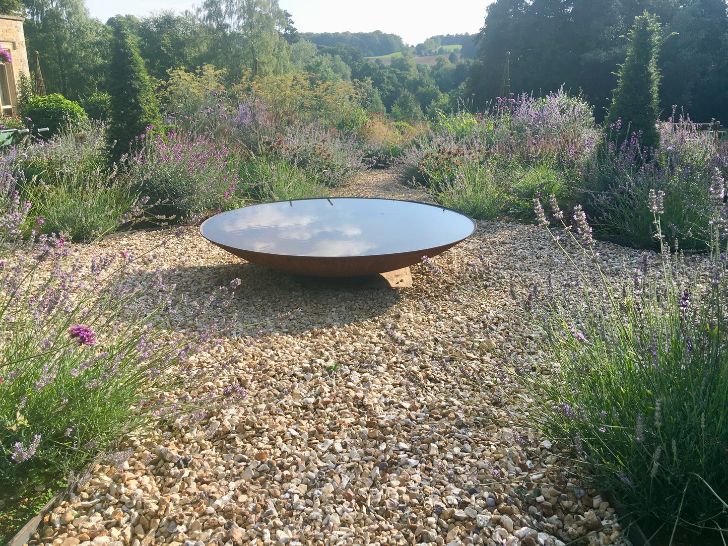 Corten steel curved round water bowl, filled with water in a garden setting with lavender and gravel, looking towards  a view over countryside