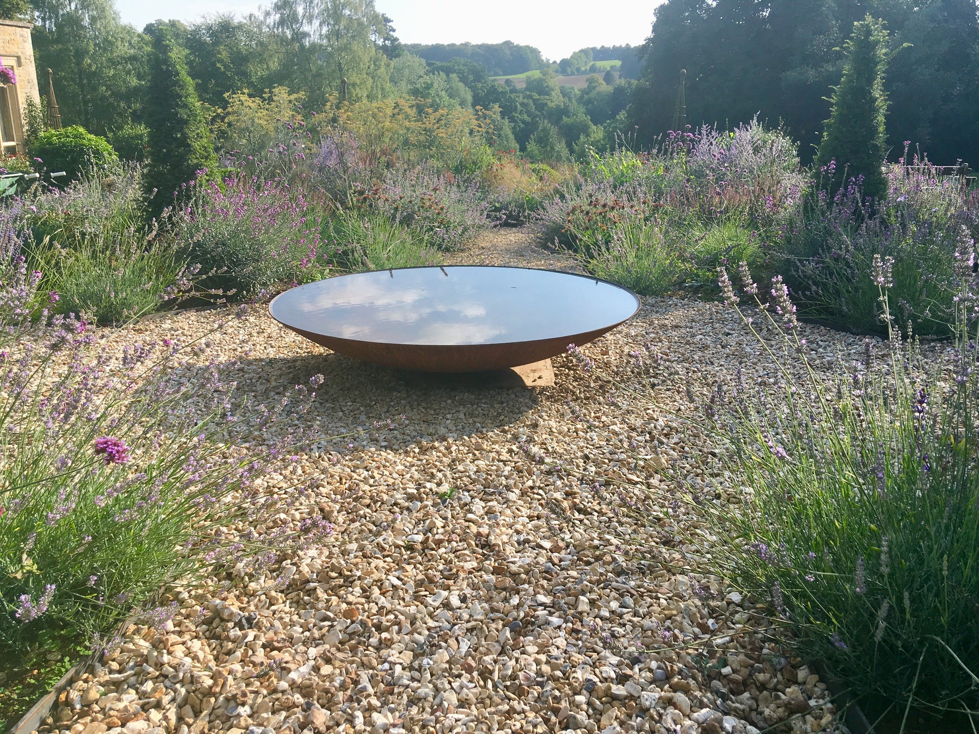 Corten steel curved round water bowl, filled with water in a garden setting with lavender and gravel, looking towards  a view over countryside