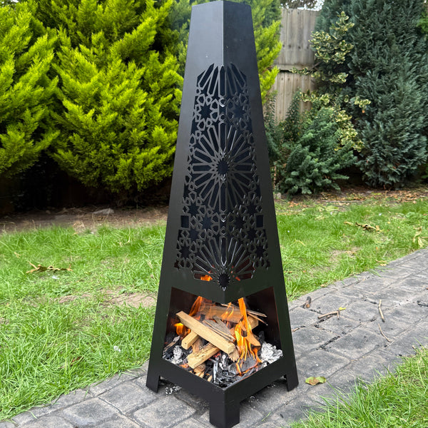 Black metal garden fire pit with decorative design on a paved pathway with greenery in the background