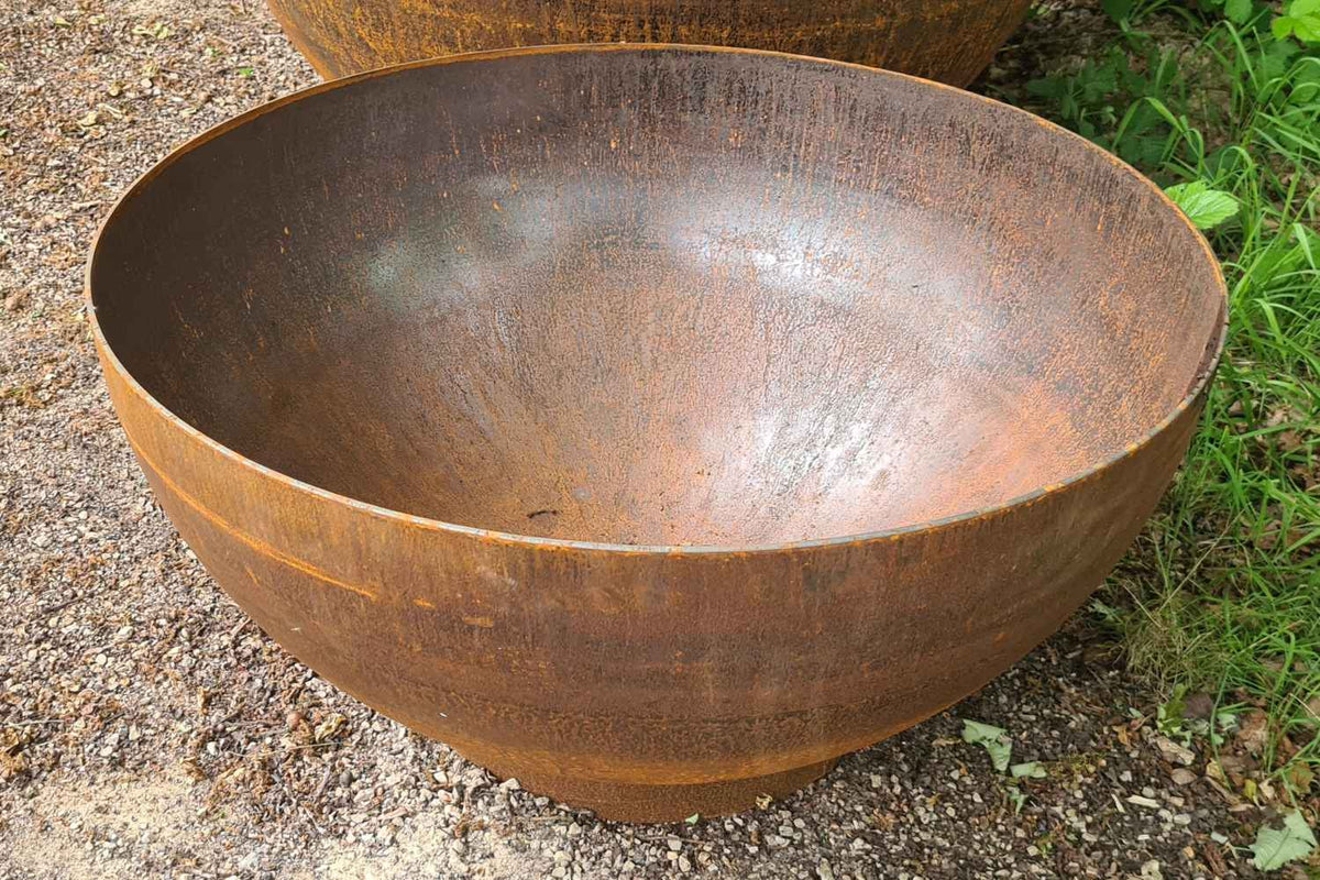 A large round deep water bowl made of weathered corten steel, showing a rustic brown and orange rust finish, set on a gravel surface with green grass visible on the right.